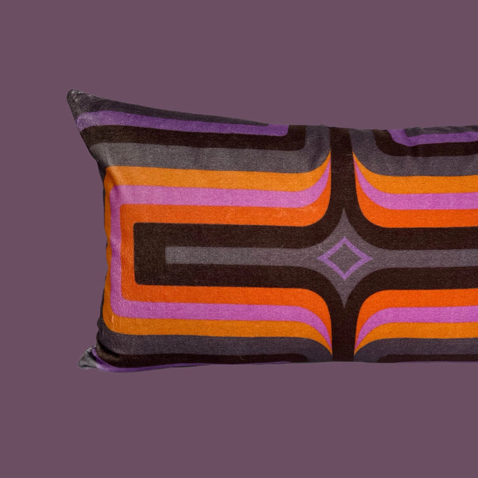 A rectangular velvet cushion with a multicolored geometric pattern, featuring stripes in shades of purple, orange, and gray.