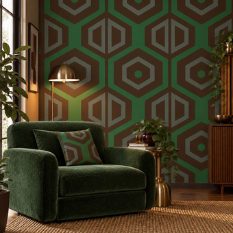 A styled interior scene featuring a deep green velvet armchair with a matching geometric throw pillow, set against a wall with green and brown geometric wallpaper. A brass floor lamp, a small side table with books and a plant, and a wooden cabinet are visible in the room.