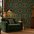 A styled interior scene featuring a deep green velvet armchair with a matching geometric throw pillow, set against a wall with green and brown geometric wallpaper. A brass floor lamp, a small side table with books and a plant, and a wooden cabinet are visible in the room.