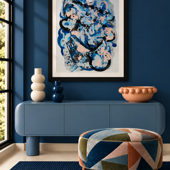 A framed abstract art print with blue, black, and white brushstrokes hangs on a dark blue wall above a blue console table. The room includes decorative vases, a terracotta bowl, a patterned blue and orange pouf, and a dark blue rug.
