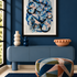 A framed abstract art print with blue, black, and white brushstrokes hangs on a dark blue wall above a blue console table. The room includes decorative vases, a terracotta bowl, a patterned blue and orange pouf, and a dark blue rug.