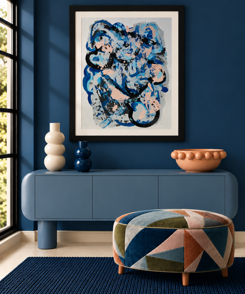 A framed abstract art print with blue, black, and white brushstrokes hangs on a dark blue wall above a blue console table. The room includes decorative vases, a terracotta bowl, a patterned blue and orange pouf, and a dark blue rug.