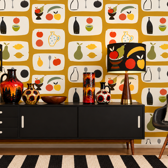 A styled interior scene featuring a bold retro wallpaper with a repeating fruit and bottle pattern on a mustard yellow background. In front of the wall is a black mid-century sideboard with two matching table lamps, decorative vases, and a black chair with a patterned pillow.