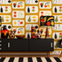 A styled interior scene featuring a bold retro wallpaper with a repeating fruit and bottle pattern on a mustard yellow background. In front of the wall is a black mid-century sideboard with two matching table lamps, decorative vases, and a black chair with a patterned pillow.