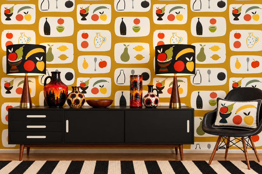 A styled interior scene featuring a bold retro wallpaper with a repeating fruit and bottle pattern on a mustard yellow background. In front of the wall is a black mid-century sideboard with two matching table lamps, decorative vases, and a black chair with a patterned pillow.