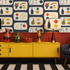 A styled interior scene featuring a bold mid-century modern wallpaper with a repeating fruit and vase pattern, a bright yellow sideboard cabinet, two matching table lamps, decorative ceramic vases, a dark blue armchair with a patterned pillow, and a black-and-white striped rug.