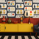 A styled interior scene featuring a bold mid-century modern wallpaper with a repeating fruit and vase pattern, a bright yellow sideboard cabinet, two matching table lamps, decorative ceramic vases, a dark blue armchair with a patterned pillow, and a black-and-white striped rug.