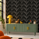 A styled interior scene showing a green ribbed sideboard with gold handles against a black wallpaper patterned with white birds. A yellow table lamp, a wooden duck figurine, a small yellow duck, colorful eggs, and a cup sit on top.