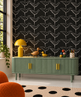 A styled interior scene showing a green ribbed sideboard with gold handles against a black wallpaper patterned with white birds. A yellow table lamp, a wooden duck figurine, a small yellow duck, colorful eggs, and a cup sit on top.