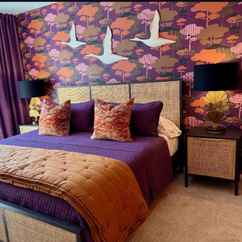 A styled bedroom featuring a bed dressed in purple bedding with a tan quilted throw, set against a bold purple, orange, and pink patterned accent wall with white bird cutouts. Purple curtains hang on the left, and two black table lamps sit on matching nightstands.