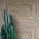 A wallpaper with a geometric diamond pattern in a tan color, displayed in a manner that mimics a woven appearance, alongside a cactus plant.